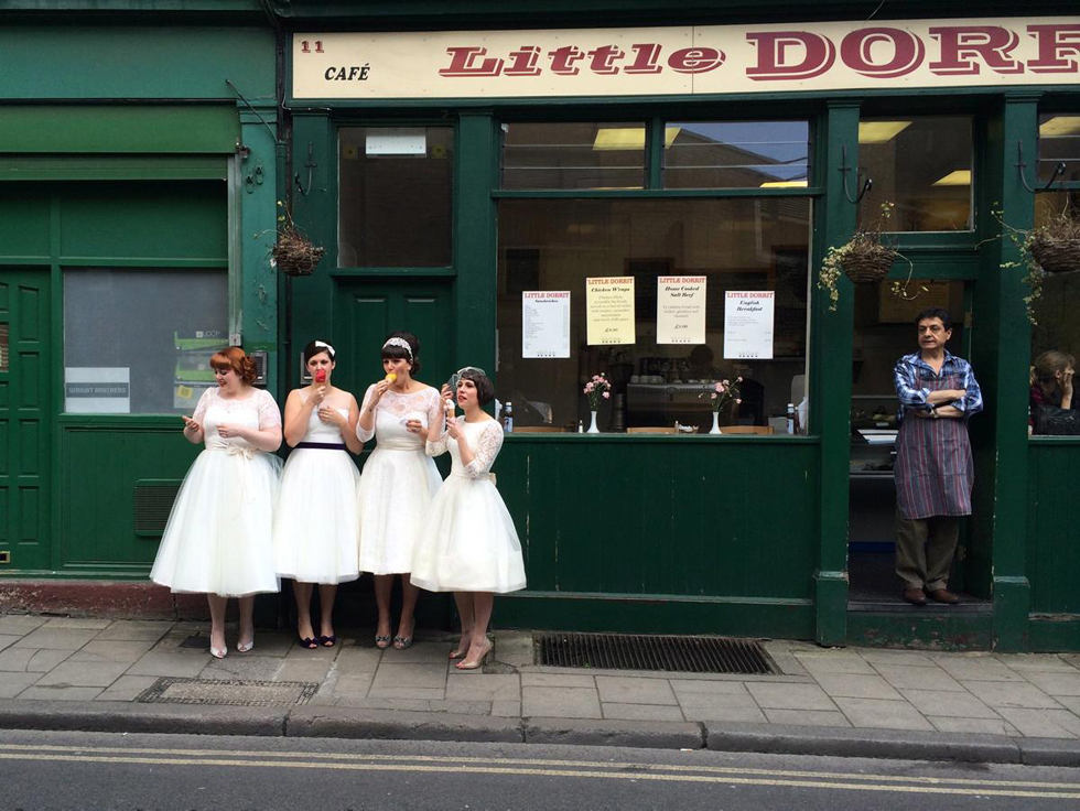 伦敦最古老的鲜活菜市场博罗集市（Borough Market），4名年轻女性身着白色礼服，站在街边吃冰淇淋，旁边的店主在看她们，摄影：Susie Stern