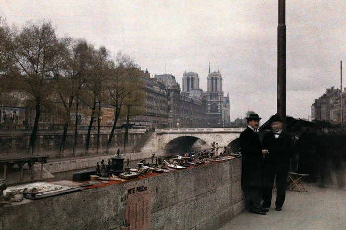 1920年代，两个男人站在塞纳-马恩省河畔待价而沽的小饰品旁边，背后就是巴黎圣母院。 PHOTOGRAPH BY JULES GERVAIS COURTELLE